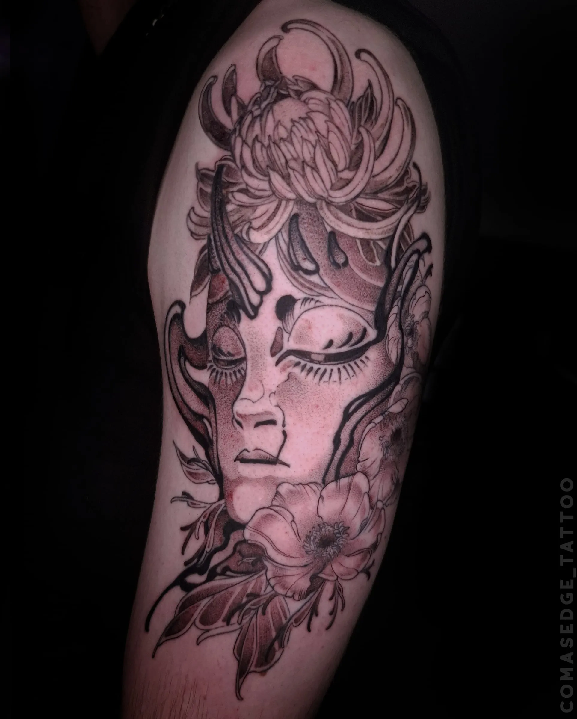 Tatouage en noir et gris d’un visage aux yeux fermés orné de fleurs et de volutes sombres, réalisé avec finesse sur le haut du bras.