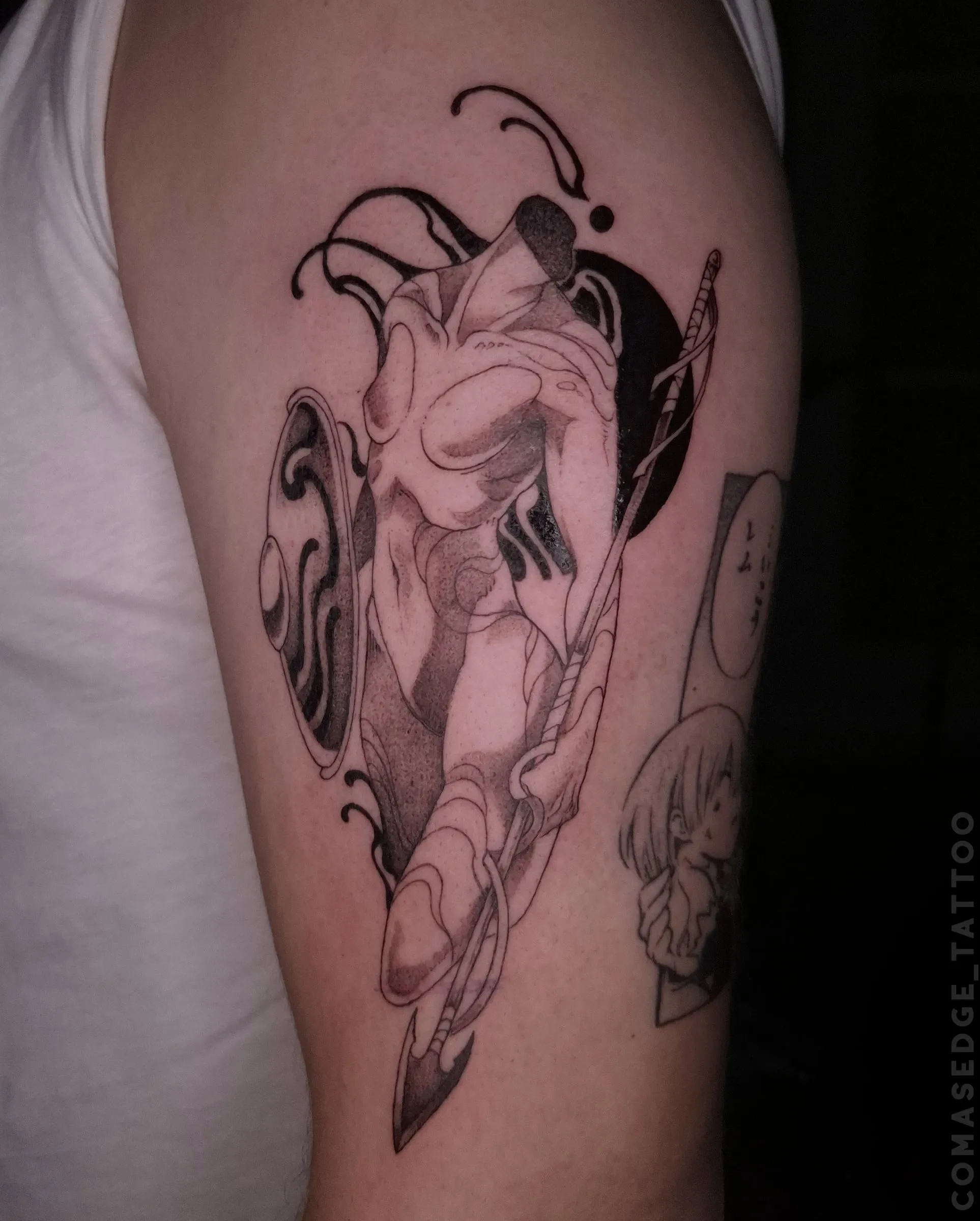 Tatouage en noir et gris d’une sculpture d’Athéna tenant une lance et un bouclier sur le haut du bras, avec des formes noires abstraites en arrière-plan.
