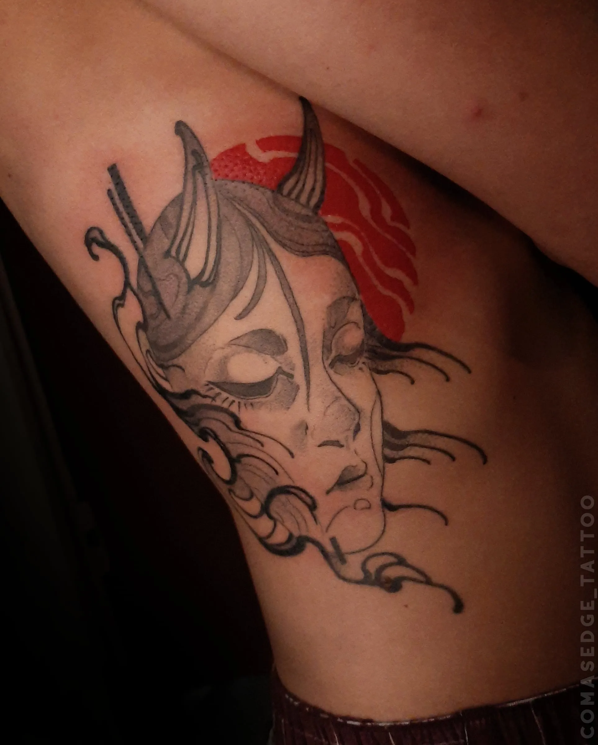 Tatouage d’un visage d’oni féminin aux yeux fermés et aux cornes apparentes sur les côtes, avec des lignes noires fluides et un disque rouge.