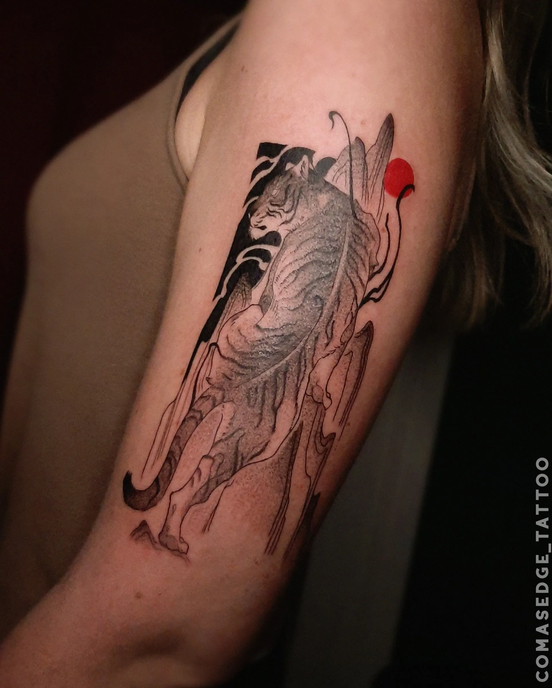 Tatouage en noir et gris d’un tigre vu de dos devant des montagnes sur le haut du bras, accompagné de formes noires et d’un cercle rouge.